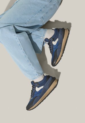 Sneakers blu in mesh e camoscio da uomo con dettagli bianchi, suola in gomma nera e tomaia testurizzata, abbinati a jeans in denim chiaro.