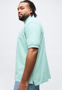 Άνδρας με κοντά dreadlocks, φορώντας φύλλο πόλο μέντας και μπλε τζιν, στέκεται σε προφίλ μπροστά σε απλό λευκό φόντο.