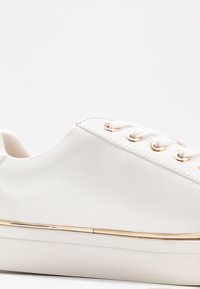 Baskets en cuir blanc avec une texture lisse, œillets dorés et semelle intermédiaire accentuée en or. Présente des lacets ronds et un design profil bas.
