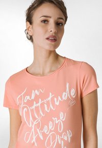 Ferskenfarvet T-shirt med korte ærmer og rund halsudskæring. Har hvid tekstgrafikdesign på forsiden. Glat stofstruktur.