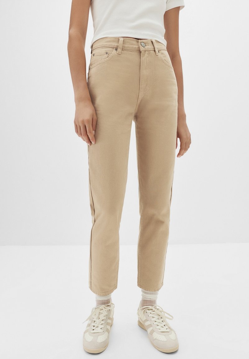 Personne portant un pantalon beige taille haute à jambes droites, des chaussettes blanches, des baskets beige et blanches, et un haut blanc à manches courtes, debout.
