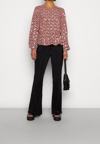 Rote und rosa gemusterte Bluse mit langen Ärmeln und elastischem Bund, kombiniert mit schwarzen gestreiften Schlaghosen und klobigen schwarzen Plateausandalen.