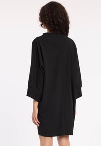 Schwarzes kurzes Kleid mit lockerer Passform, Dreiviertelärmeln und geradem Saum. Hergestellt aus glattem Material mit minimalistischem Design.