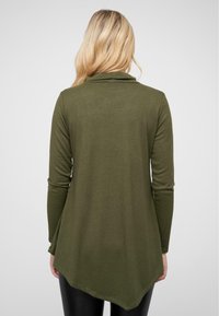 Tunique à manches longues vert olive avec col drapé, présentant un ourlet arrondi et un tissu texturé, associée à des leggings noirs.