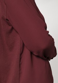 Prenda burdeos con tejido suave y texturizado con un corte holgado. Manga larga y drapeado sutil en el lateral.