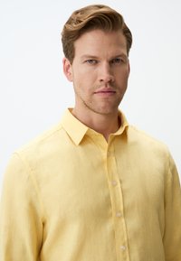 Camicia in lino giallo chiaro con colletto classico, chiusura frontale con bottoni e un aspetto tessuto, caratterizzata da un leggero luccichio e cuciture minime.