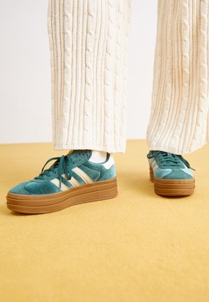 Des baskets en suède teal présentent une épaisse semelle plateforme marron, des rayures blanches et des lacets assortis, complétées par un pantalon en tricot ivoire texturé.