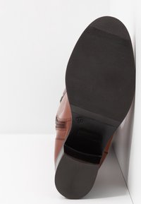 Semelle d'une chaussure en cuir marron avec talon et bout en caoutchouc noir, taille 37 visible sur la semelle, chaussure posée à la verticale contre une surface blanche.