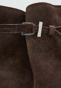 Material de ante marrón con una textura suave, que presenta una hebilla rectangular plateada y un detalle de correa a juego.