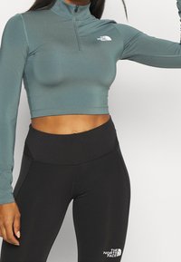 Helles taubenblaues, kurz geschnittenes Langarmoberteil mit einem halben Reißverschluss-Kragen, mit weißen Logo-Details. Kombiniert mit hoch taillierten schwarzen Leggings.