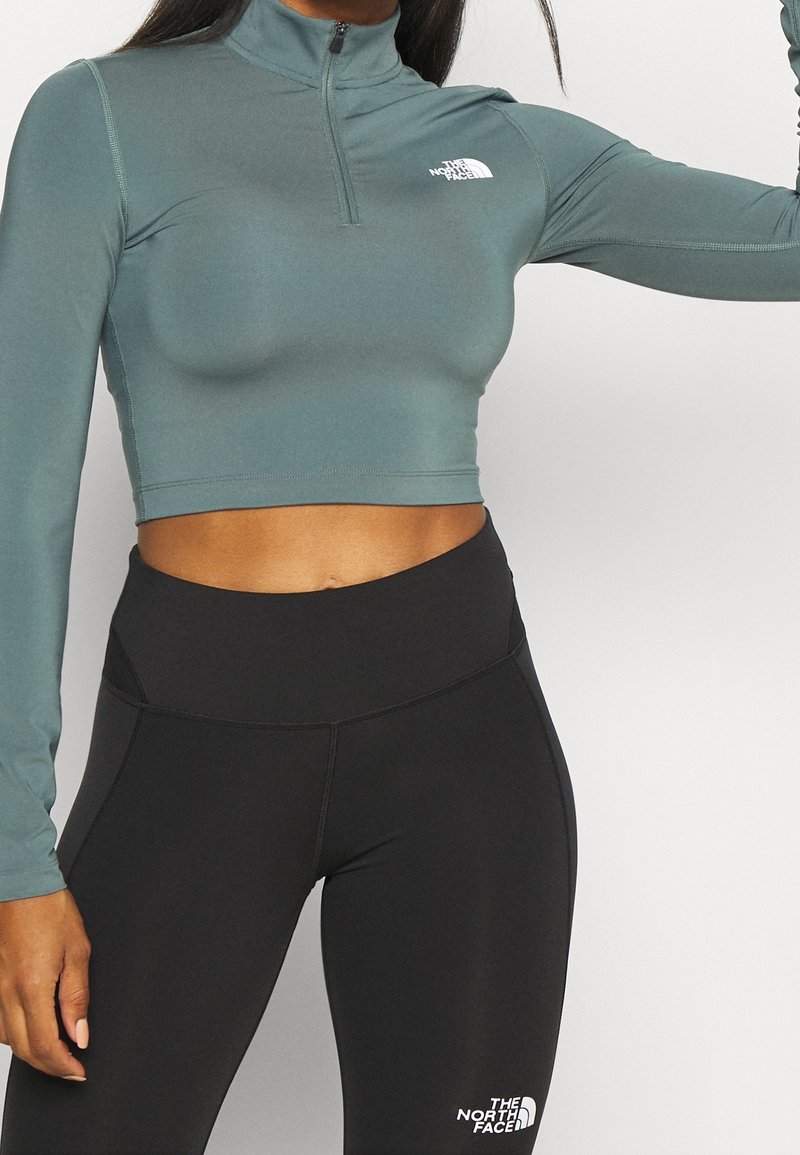 Helles taubenblaues, kurz geschnittenes Langarmoberteil mit einem halben Reißverschluss-Kragen, mit weißen Logo-Details. Kombiniert mit hoch taillierten schwarzen Leggings.