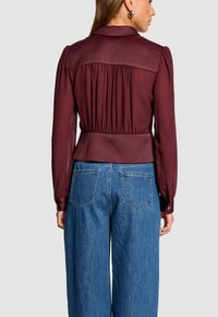 Blusa borgogna con colletto, schiena arricciata e maniche lunghe a sbuffo. Abbinata a jeans in denim blu a gamba larga con tasche posteriori.