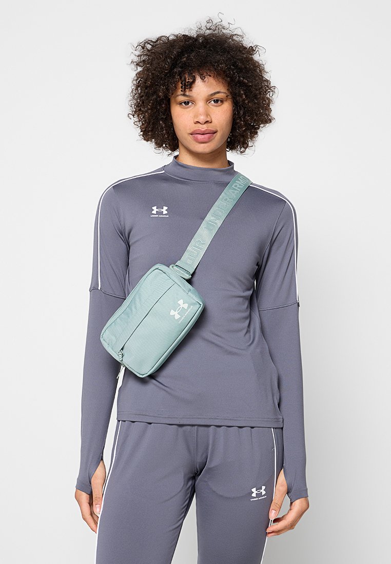 Mujer con cabello rizado que lleva un chándal gris de Under Armour y un bolso bandolera verde claro con el logo de Under Armour.