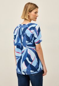 Blusa blu a maniche corte con un motivo a foglia in diverse tonalità di blu, accentuata da linee rosa e bianche. Tessuto morbido e leggero.