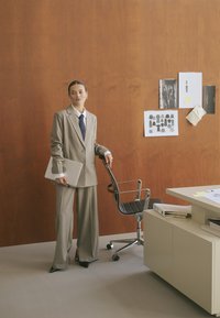 Costume gris sur mesure avec un pantalon large et une veste, associé à une chemise blanche et une cravate foncée ; tenant un ordinateur portable dans un cadre de bureau.