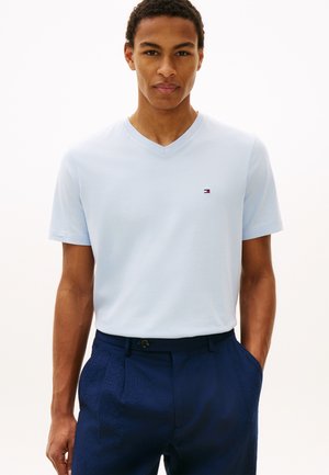 Άνδρας που φοράει ανοιχτό μπλε T-shirt με V λαιμόκοψη και μικρό λογότυπο, μαζί με σκούρο μπλε παντελόνι, στέκεται με το ένα χέρι στην τσέπη μπροστά σε λευκό φόντο.