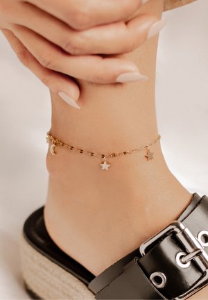 Goldene Fußkette mit Mond- und Sternanhängern an einem nackten Knöchel. Sie hat eine gedrehte Gliederkette und einen dezenten Glanz vor einem hellen Hintergrund.