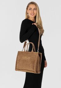 Bruine fleece handtas met een rechthoekige vorm, voorzien van twee handvatten en een afneembare schouderband. Gouden "REPLAY" logo aan de voorkant.