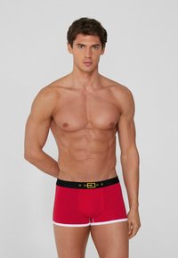 Červené boxerky s kontrastním bílým lemem a černým pasem s detailem zlaté spony. Hladká textura, přiléhavý design.