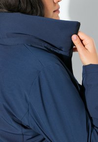 Žena v námořnickém modrém saku, která si upravuje zip u límce, na jednobarevném světlém pozadí.