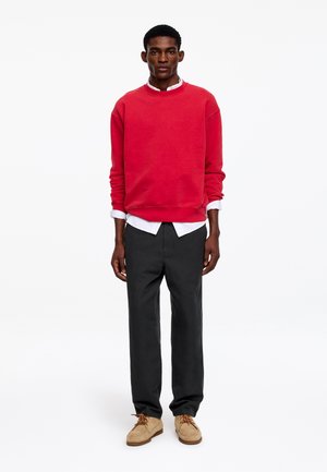 Jeune homme debout, portant un sweat-shirt rouge sur une chemise blanche, un pantalon noir et des chaussures beiges, sur un fond blanc uni.