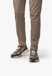 Zapatillas marrones y grises con materiales de malla y ante, diseño en capas, acentos contrastantes y suela texturizada. Llevadas con pantalones beige.