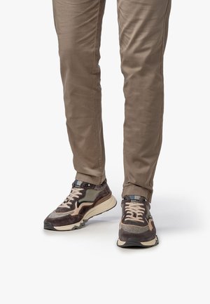 Baskets marron et gris avec des matériaux en mesh et en suède, design en couches, accents contrastants et semelle texturée. Portées avec un pantalon beige.