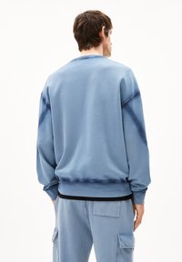 Man die met zijn rug naar de camera staat en een losse blauwe sweatshirt en bijpassende cargo broek met zakken draagt, tegen een eenvoudig witte achtergrond.