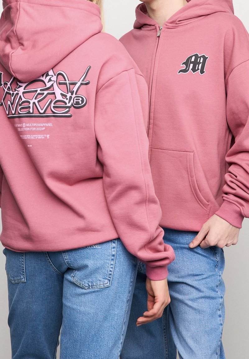 Sudaderas rosas con cremallera frontal, que presentan un logo en el pecho y un estampado gráfico en la parte posterior. Fabricadas en algodón con una textura suave.