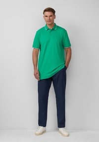 Groene polo met witte accenten, gecombineerd met donker marineblauwe broek. Model staat casual met handen in de zakken, voor een effen achtergrond.