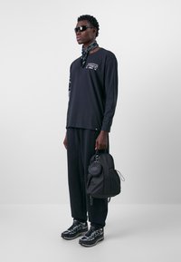 Langärmliges schwarzes Shirt mit grafischen Details, schwarze Hose, schwarzer Rucksack, schwarze Sonnenbrille und gemusterte Sneakers mit grauen Akzenten.