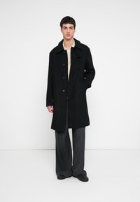 Zwarte wollen jas met een kraag, knoopsluiting aan de voorkant en zijzakken. Gestyled over een lichtgekleurde top en gecombineerd met losse grijze broek.