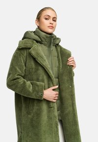 Groene oversized schapenvachtjas met hoge kraag, gelaagd over een groene pufferjack. Heeft een knoopsluiting bij de mouwen en zakken aan de zijkant.
