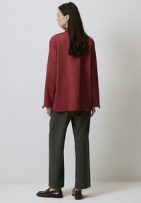 Blusa bordeaux lunga con una vestibilità rilassata, maniche lunghe e dettagli sui polsini con bottoni, abbinata a pantaloni larghi grigi e scarpe scure.