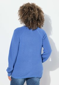 Modrý pletený svetr s ribbovanou texturou, raglánovými rukávy a zaobleným lemem. Nosený s světlými džínami, které zdůrazňují ležérní styl.