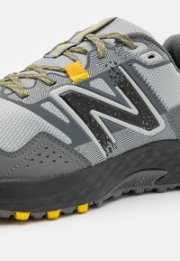 Šedá sportovní bota s geometrickým vzorem. Obsahuje žluté akcenty, černé logo a strukturovaný svršek. Černá gumová podrážka s žlutými trakčními hroty.