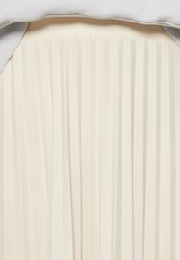 Tela plisada de color crema con una textura suave y lisa. La parte superior presenta una banda de color gris claro, que realza el diseño de la prenda.