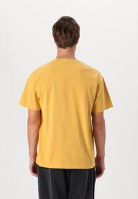 T-shirt gialla in cotone con collo arrotondato, maniche corte e vestibilità rilassata, vista da dietro. Texture liscia, senza modelli visibili.