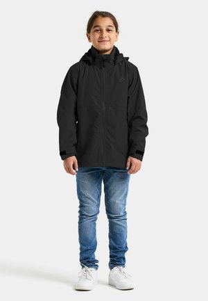 Zwarte waterdichte jas met capuchon, voorzien van ritssluiting, elastische manchetten en een gladde textuur. Gecombineerd met blauwe spijkerbroek en witte sneakers.