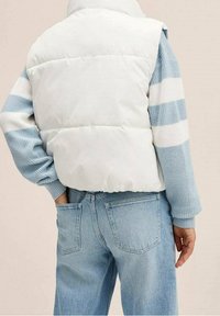 Lättvadderad väst i vitt med hög krage, kombinerad med en blå och vit randig stickad tröja och ljusblå denimjeans.