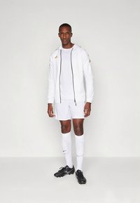 Bílý sportovní teplákový komplet se zipovou bundou a odpovídajícími šortkami. Černé a zlaté logo. Černé kopačky a bílé vysoké ponožky až po kolena.