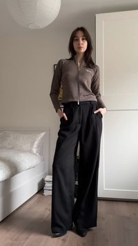 Brown zip-up cardigan with ribbed texture, paired with high-waisted black wide-leg trousers. Background features a bed and storage.