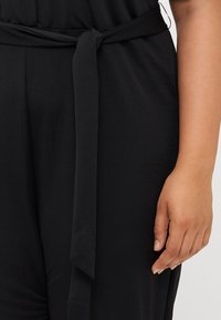 Close-up of a hand holding a black jumpsuit with a tied waist. The fabric appears soft and smooth, emphasizing the garment's detail.