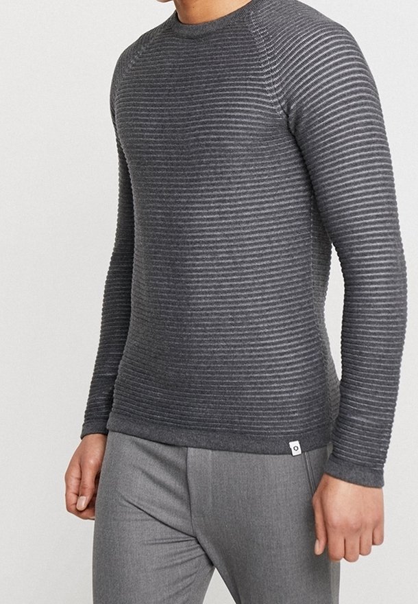 Homme portant un pull côtelé gris foncé ajusté à manches longues et un pantalon gris clair sur un fond clair uni.