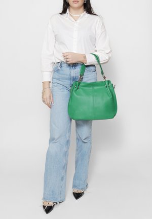 Borsa a mano in pelle verde con forma arrotondata, manici superiori e ferrature argentate. Il modello indossa una camicia bianca e jeans azzurri.