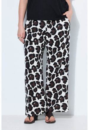 Person iført bukser med brede ben med sort og mørkebrunt leopardmønster, sort ærmeløs top og sorte åbne sandaler, stående mod en hvid væg.