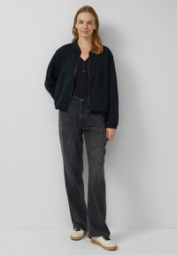 Czarna kurtka typu bomber zapinana na zamek z przodu, w połączeniu z ciemnoszarymi dżinsami i beżowymi trampkami. Charakteryzuje się luźnym krojem i prostym designem.