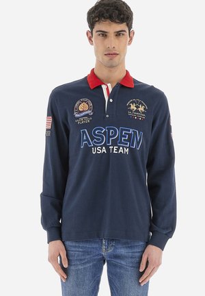 Polo a maniche lunghe blu navy con colletto rosso, loghi ricamati e lettere bianche. Presenta una vestibilità comoda e polsini a coste.