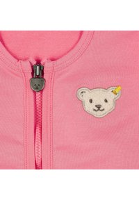 Maglietta rosa con zip, caratterizzata da un'applicazione a forma di faccia d'orso sul lato sinistro e un tirante a forma di orso. Tessuto di cotone morbido e liscio.