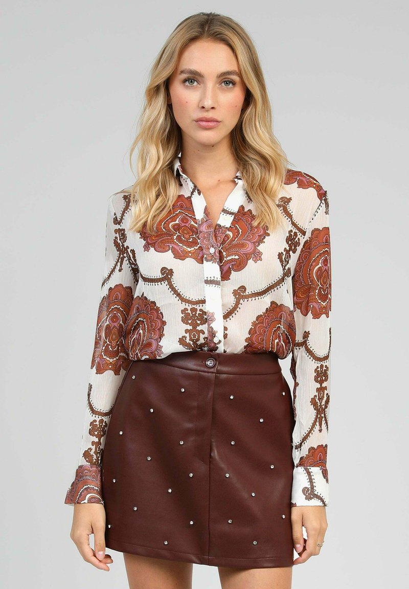 Blouse à manches longues avec des motifs floraux en rouge et orange sur un fond blanc, associée à une jupe en simili cuir marron avec des clous argentés.